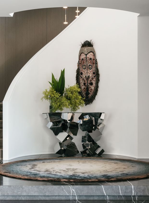 A refined contemporary entry foyer featuring a sculptural white curved wall, dark timber wall panelling, pendant lighting, a large carved decorative mask, lush indoor plants, and a faceted black-and-silver console table. A large muted rug sits above polished dark stone steps with white veining.