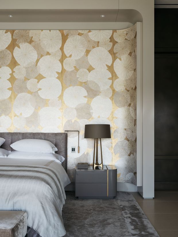 A refined bedroom interior featuring a large upholstered bed with layered white and grey bedding, a dramatic botanical-patterned feature wall in soft beige, cream and white, and a dark contemporary bedside table with a sculptural floor lamp. Grey carpeting, curved ceiling detailing and dark built-in cabinetry complete the sophisticated scheme.