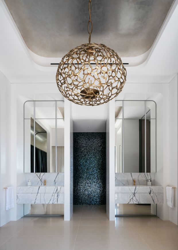 Symmetrical, high-end bathroom interior with twin floating marble vanities, integrated sinks, large mirrored panels, and a dark textured feature wall in the centre. A sculptural brass-gold spherical pendant hangs beneath a recessed, curved ceiling detail. The room uses white walls, pale stone flooring, veined white marble, and reflective surfaces for a refined contemporary look.