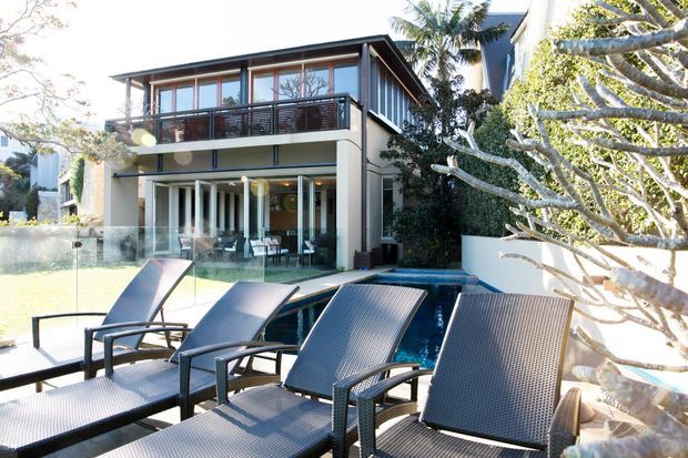 A contemporary two-storey residence viewed from a poolside terrace, featuring extensive glazing, a covered ground-floor outdoor living area, an upper balcony with dark balustrade, and a low-pitched roof. Several dark woven sun loungers sit beside the narrow swimming pool, with mature tropical and sculptural planting around the property.