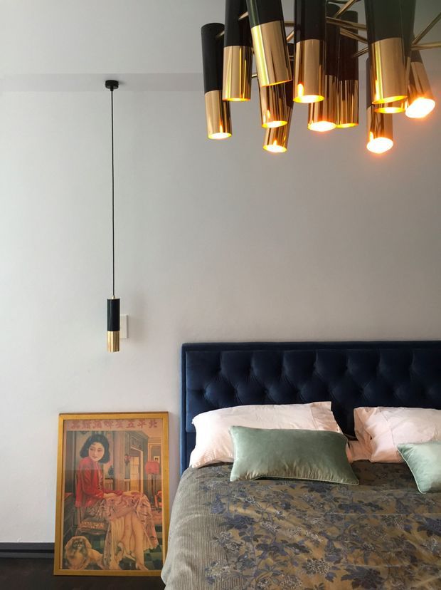 A stylish bedroom with a dark navy-blue tufted upholstered bedhead, patterned bedding, and pale green cushions. The room has a neutral light wall, dark flooring, a framed decorative artwork leaning against the wall, and distinctive hanging pendant lights in black and metallic brass/gold finishes, including a clustered multi-light ceiling fixture and a single bedside pendant.