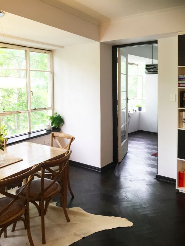 Bright dining area with a timber dining table and bentwood chairs beside large windows. The room has white walls, dark herringbone timber flooring, black skirting boards and window frames, and built-in open shelving. A glazed doorway leads to a bright adjoining space, while a neutral hide rug defines the dining zone.