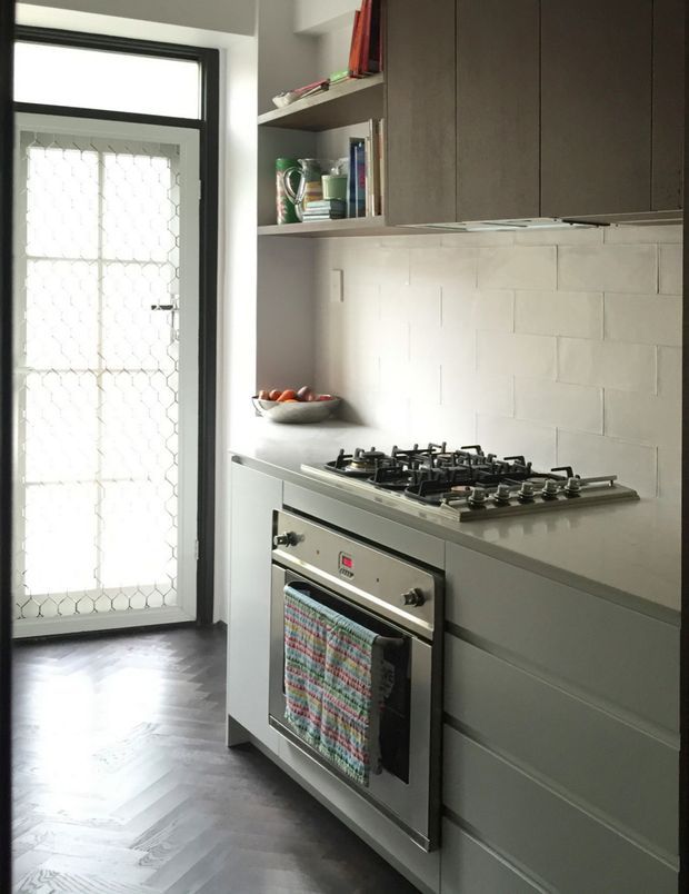 Compact contemporary kitchen featuring dark grey built-in cabinetry, a pale stone or solid-surface benchtop, white rectangular tiled splashback, stainless-steel oven and gas cooktop, and open shelving. A black-framed glazed door with security mesh provides natural light, while the dark timber herringbone floor adds contrast.