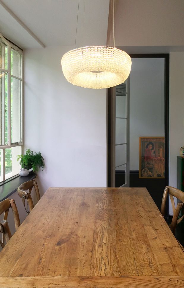 A bright dining room centred on a large rustic timber dining table with matching wooden chairs. Tall multi-pane windows line the left side, bringing in abundant natural light and views of greenery. A prominent woven or beaded pendant light hangs above the table, while a dark-framed glazed doorway opens to an adjacent room displaying artwork. The room has white walls, a pale ceiling, and a warm, natural material palette.