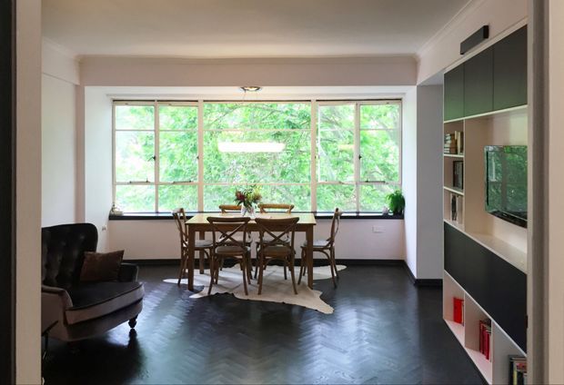 A refined dining and living area viewed from within the room. A timber dining table and chairs sit in front of large multi-pane windows overlooking leafy greenery, while a dark leather armchair occupies the foreground. The room features dark herringbone timber flooring, pale walls, and a full-height built-in entertainment and shelving unit with dark cabinetry and a recessed television niche. The atmosphere is warm, calm, and sophisticated.