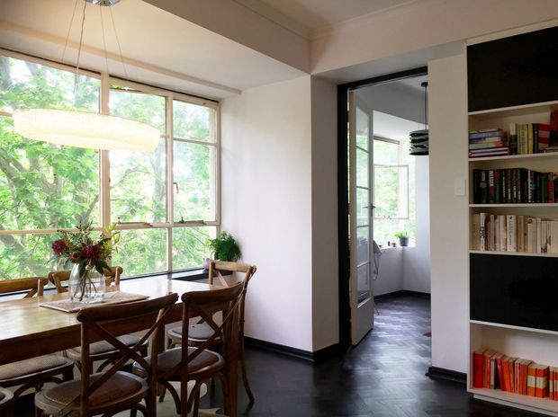 A warm, refined dining room with a timber dining table and cross-back chairs positioned beside large timber-framed windows overlooking leafy greenery. A sculptural pendant light hangs above the table, while a dark herringbone timber floor and built-in open shelving filled with books define the room. A black-framed glazed doorway leads to a bright adjoining space.