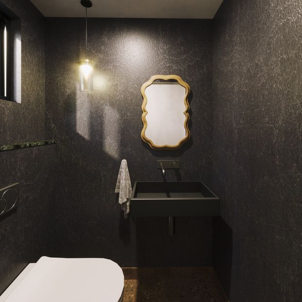 A compact, dark powder room with textured charcoal walls, a black wall-mounted rectangular basin and vanity, a decorative gold-framed scalloped mirror, and a pendant light. A partial white toilet is visible in the foreground, with a small window and dark stone-like surfaces adding contrast. The space has a dramatic, moody, luxurious feel.