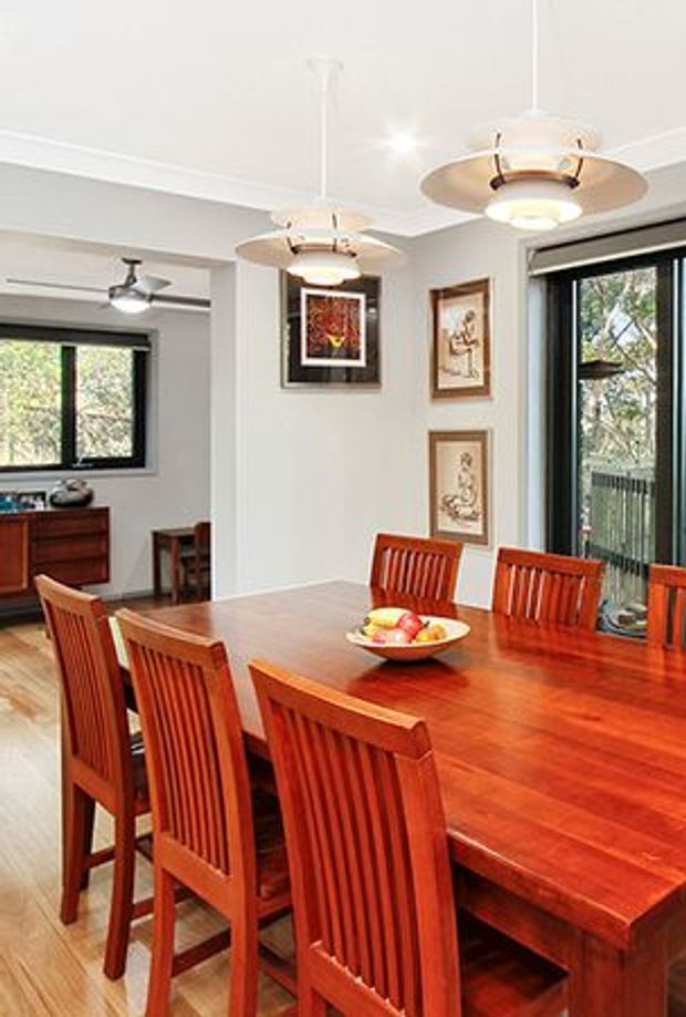 A warm, traditional dining room dominated by a large polished reddish timber dining table with matching slat-back chairs. The room has light painted walls, timber flooring, framed artwork, pendant lights, and large black-framed windows and glazing overlooking greenery. An adjacent sitting or living area is visible through the opening at left.