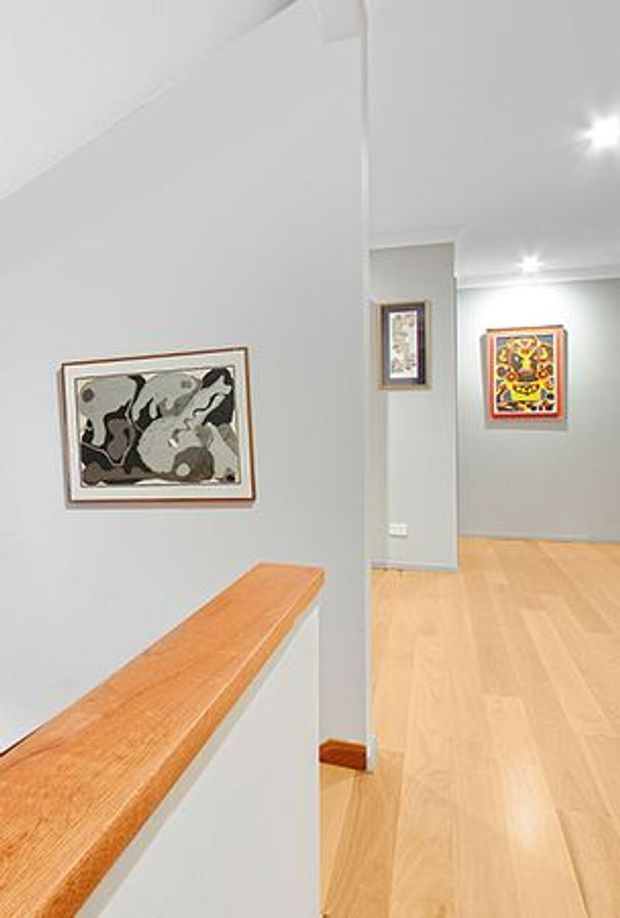 A bright minimalist interior hallway or landing with pale grey walls, light timber flooring, and a white half-height stair or balustrade wall capped with a warm timber handrail. Framed artwork decorates the walls, and recessed ceiling lights provide even illumination. The space opens toward another gallery-like corridor in the background.