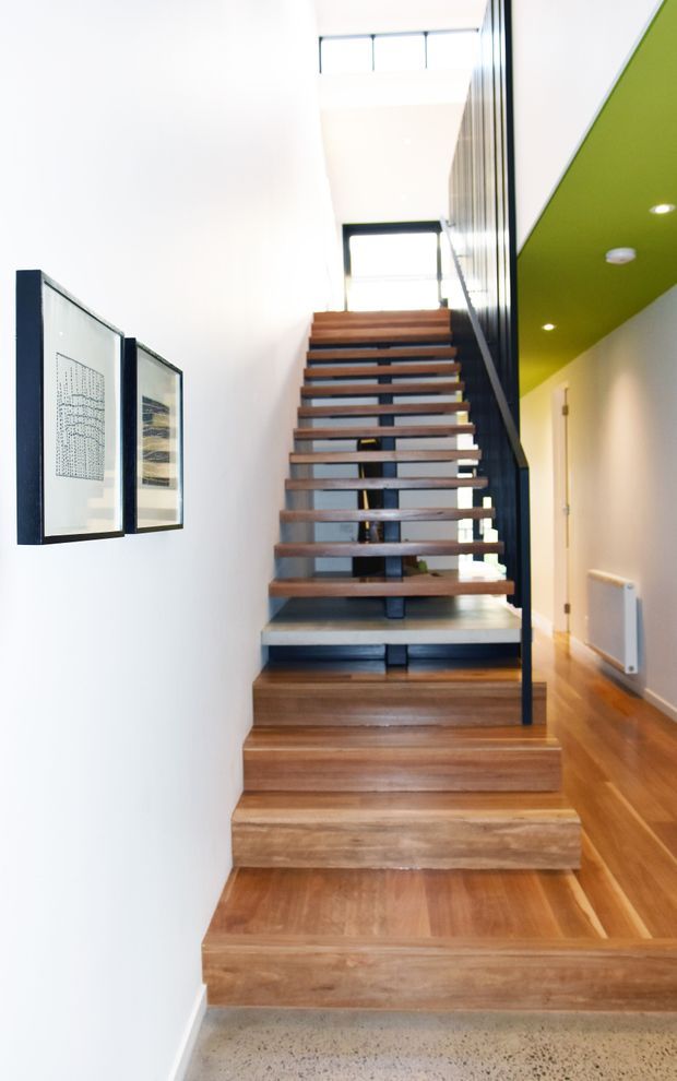 Bright contemporary interior hallway focused on a staircase with broad timber treads, open risers, and a dark metal balustrade. White walls display framed artwork, while a vivid green ceiling plane with recessed downlights accents the upper-right corridor. Timber flooring continues along the hallway, and the stair rises toward a high glazed landing.