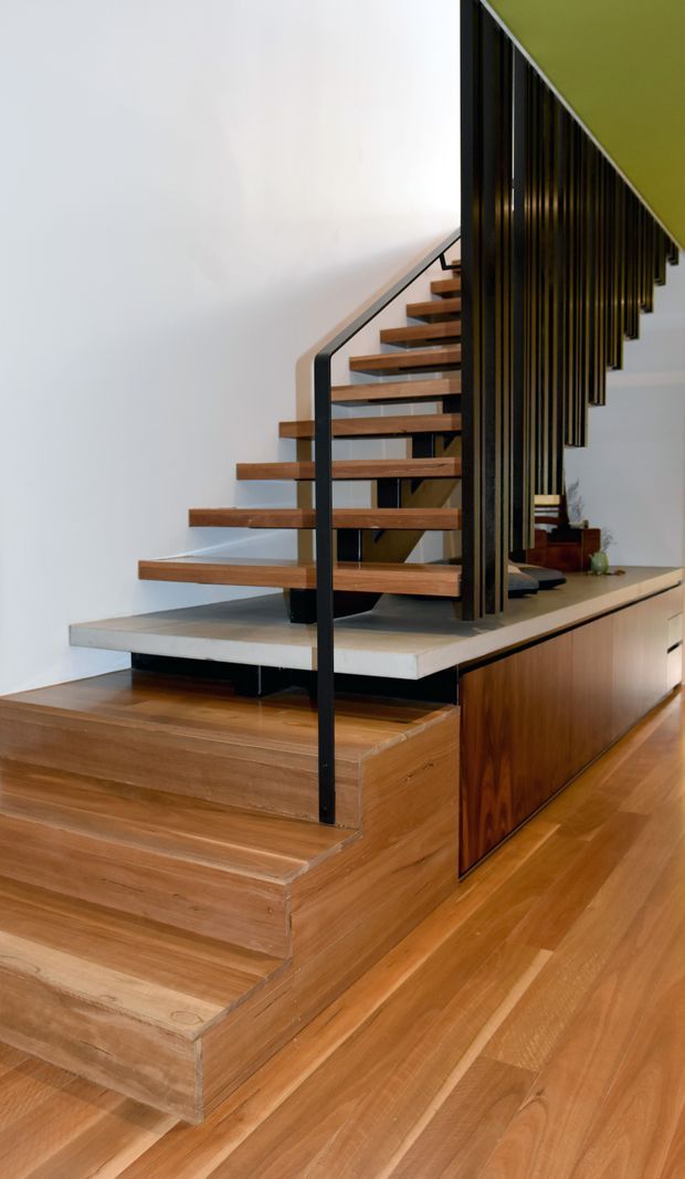 Interior view of a contemporary stairway with floating timber treads, black metal handrails, and a dramatic vertical black slat screen forming a balustrade. The stair structure rises from a raised timber platform with integrated cabinetry, while warm timber flooring continues through the surrounding space. White walls and a lime-green ceiling accent create a strong modern contrast.