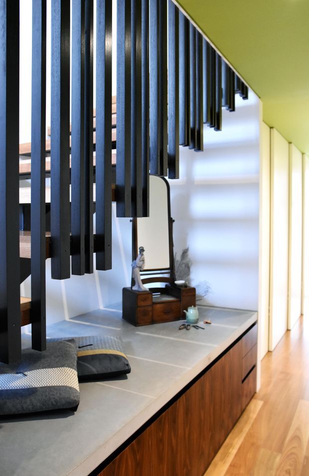 Interior view of a contemporary stairway and hallway. A sculptural staircase with numerous dark vertical timber or metal slats descends beside a long built-in timber cabinet topped with a pale grey surface. The space has white walls, warm timber flooring, and a small vintage-style console with mirror beneath the stair structure. Soft daylight enters from the right, creating a refined, modern architectural feature.