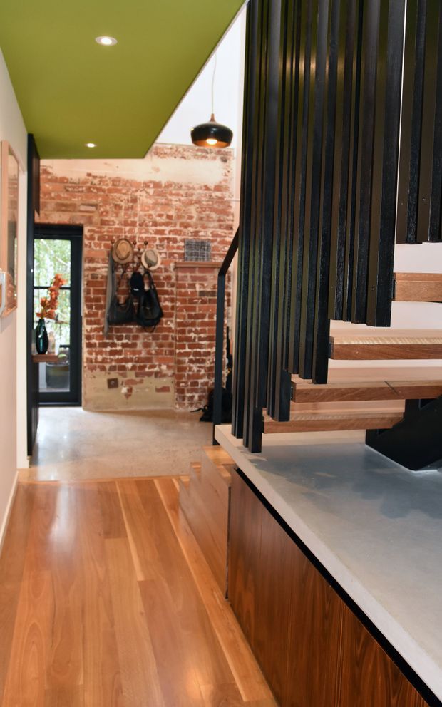 Interior view of a stylish industrial-transitional home, looking past a timber staircase with black vertical slatted balustrade toward an exposed red-brick wall and glazed exterior door. Warm timber flooring and stair treads contrast with dark built-in cabinetry and a light grey benchtop in the foreground. A green painted ceiling, pendant light, wall hooks, and recessed lighting add distinctive interior character.