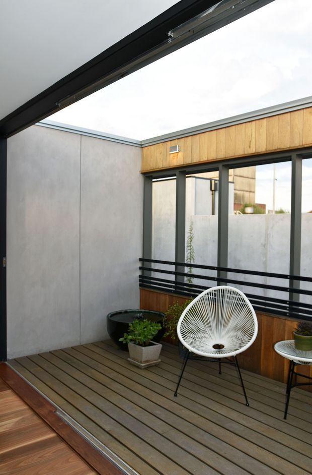 A compact open-air courtyard terrace with timber decking, smooth grey rendered walls, warm timber wall cladding, black-framed glazing and horizontal black railings. Potted plants and a white woven lounge chair create a relaxed outdoor seating area beneath a large retractable or open roof aperture. The space has a clean, contemporary urban character.