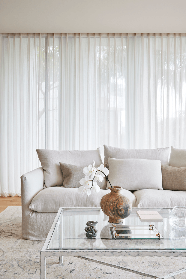 Bright, softly styled living room with a large cream sectional sofa layered with neutral cushions, floor-to-ceiling sheer curtains, a pale patterned rug, and a clear glass coffee table displaying a rustic ceramic vessel, orchid arrangement, decorative sculpture, books, and glassware. The room has a calm, refined neutral palette with abundant diffused daylight and views of trees through the curtains.