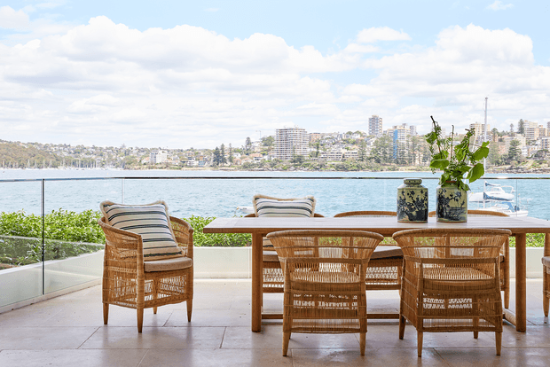 Bright waterfront outdoor dining terrace with a timber dining table and woven rattan chairs, overlooking a calm harbour and urban shoreline. A glass balustrade, pale tiled flooring, striped cushions, and potted greenery create an airy coastal setting.