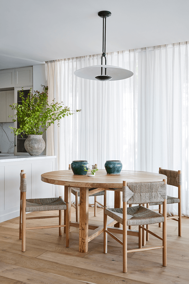 Bright, refined dining area with a round light-oak timber dining table and woven upholstered dining chairs. Full-height sheer curtains create a softly illuminated backdrop, while white built-in cabinetry and a marble-look kitchen surface sit to the side. A sculptural white pendant light hangs above the table, with ceramic vessels and a large textured planter adding restrained decorative accents.