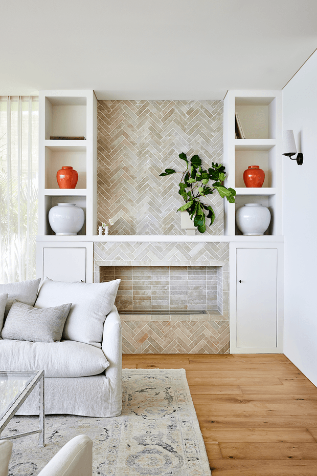 Bright, refined living room featuring a white built-in fireplace wall with symmetrical open shelving and lower cupboards. The fireplace surround and interior are clad in warm beige and light grey herringbone-patterned tiles, complemented by light timber flooring, a pale upholstered sofa, neutral rug, decorative pottery, and a leafy plant. Sheer curtains and a minimalist wall sconce add a soft, airy feel.