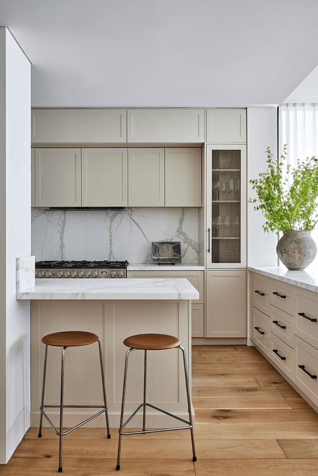 A refined, light-toned kitchen with cream shaker-style cabinetry, a marble-look stone backsplash and island benchtop, integrated storage, a stainless-steel range cooker, and two brown leather bar stools. Warm timber flooring contrasts with the pale cabinetry, while a large potted tree sits beside the bright window.