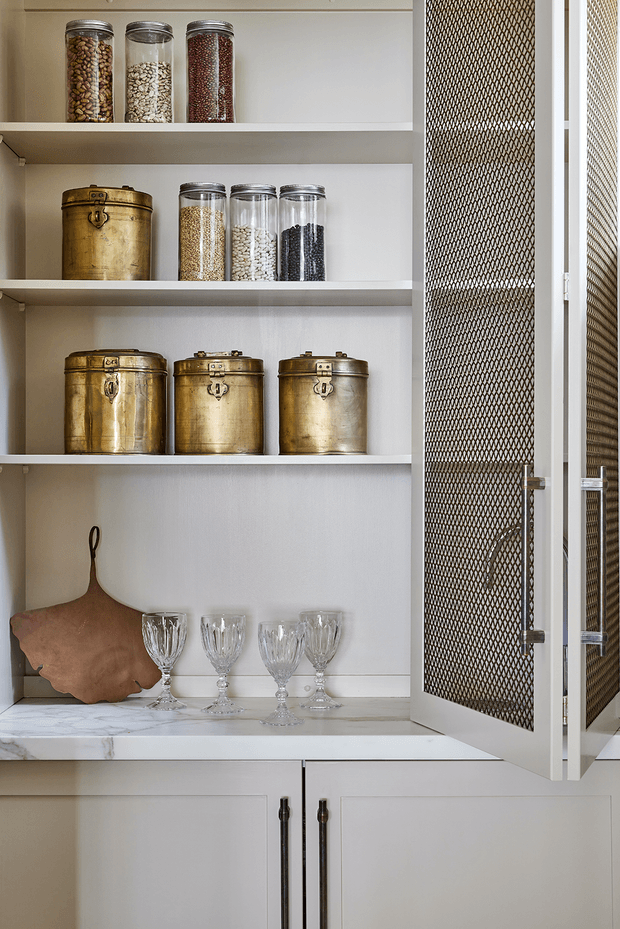 A built-in pantry or storage cupboard with open white shelving displaying glass jars of dry goods, brass canisters, and crystal glasses. The lower section has white cabinetry with dark metal handles, while a hinged mesh-front door adds ventilation and visual texture. A pale marble countertop and warm brass accessories create a refined, traditional look.