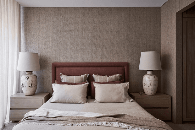 A softly lit, symmetrical bedroom with a textured taupe wall, upholstered muted-rose headboard, layered neutral bedding, matching wooden bedside tables, and ceramic table lamps. Sheer curtains provide gentle daylight on the left, while a dark timber doorway frames the right side.