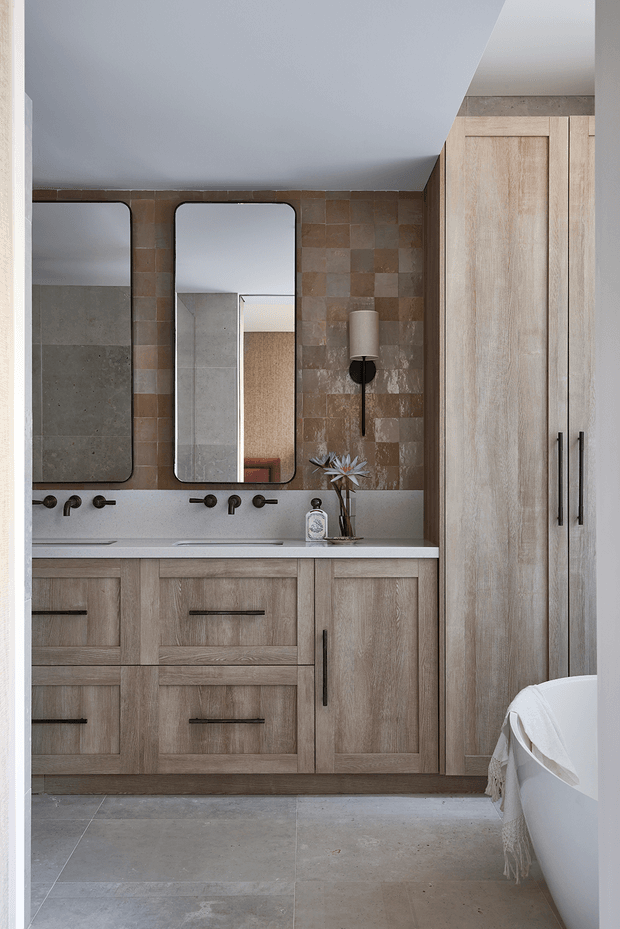 A refined bathroom vanity area with a long double-sink cabinet, two rounded-rectangle mirrors, wall-mounted sconces, and full-height light timber cabinetry. The space features a warm brown and taupe patchwork tile backsplash, pale stone-look benchtop, large grey-beige floor tiles, black tapware and hardware, and part of a freestanding white bathtub visible at right.