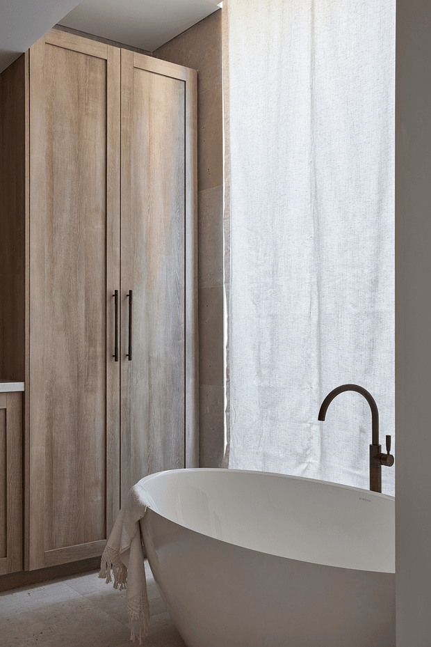 A softly lit contemporary bathroom featuring a freestanding white bathtub in the foreground, a dark bronze curved wall-mounted faucet, tall light timber cabinetry with black handles, large beige stone-look wall tiles, and a sheer white linen curtain filtering daylight.