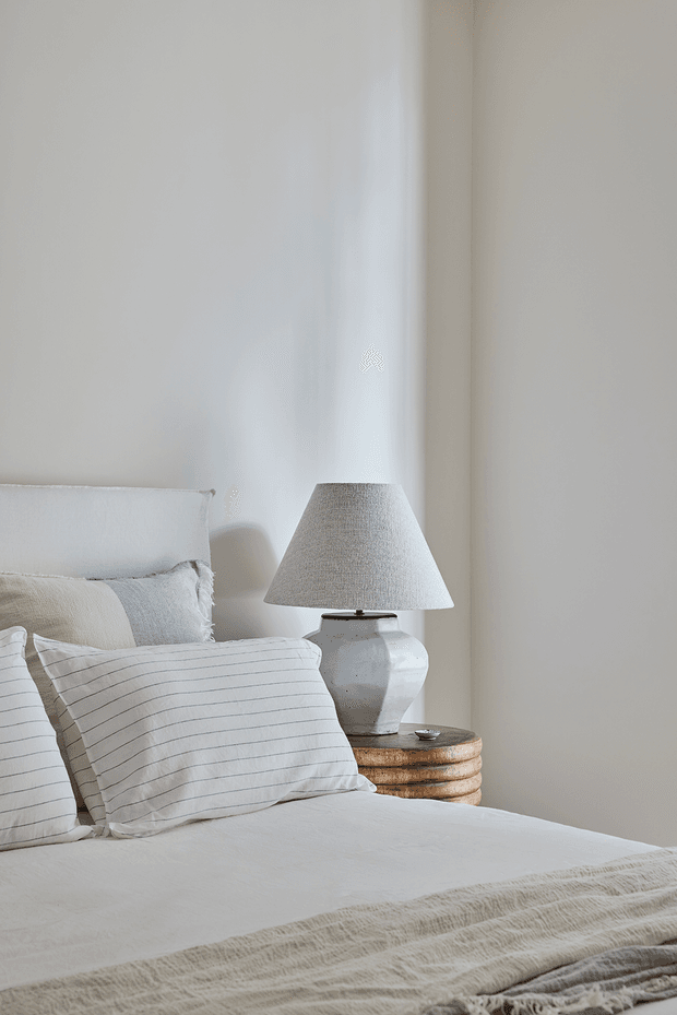 A softly lit, neutral-toned bedroom vignette featuring a neatly made bed with layered white and beige bedding, striped linen pillows, a pale upholstered headboard, and a ceramic table lamp on a rounded wooden bedside table. The overall styling is calm, natural, and understated.