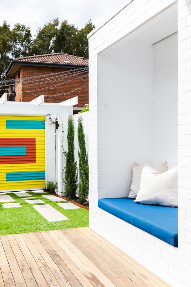 A bright contemporary outdoor courtyard featuring a white-painted brick structure with a recessed built-in window seat upholstered in vivid blue and layered with white cushions. A timber deck leads to a small artificial-grass lawn with rectangular stepping stones, tall narrow plants, and a colourful striped yellow, red, and turquoise garage or boundary door. A neighbouring brick building with a tiled roof and overhead festoon lights is visible in the background.