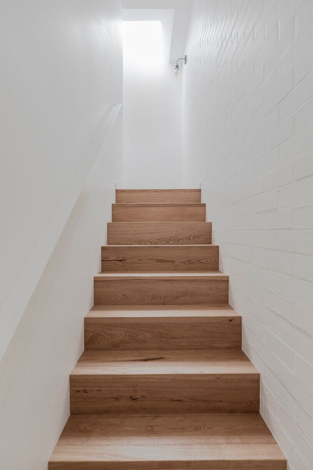 A narrow, minimalist stairway viewed from below, with broad natural timber treads and risers between smooth white walls. The right wall has painted white brickwork, while a small skylight or roof opening brightens the upper landing. A simple exposed wall light adds a subtle architectural detail.