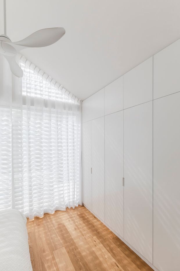 Bright, compact minimalist bedroom with floor-to-ceiling white built-in wardrobes, pale timber flooring, sheer white curtains, and a sculptural white ceiling fan. The edge of a bed is visible at lower left, while abundant filtered daylight creates an airy, calm atmosphere.