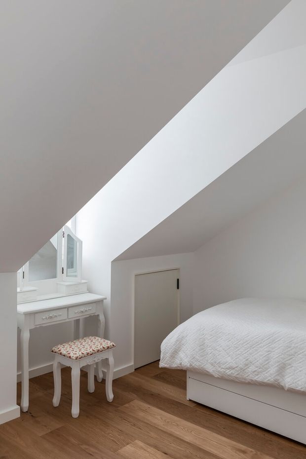 Bright, compact attic bedroom with a sharply sloped white ceiling and matching white walls. The room contains a white bed with quilted bedding, a small ornate white dressing table and upholstered stool, a mirrored dressing area, a simple white door, and warm timber flooring. The minimalist furnishings and abundant white surfaces create a clean, airy atmosphere.