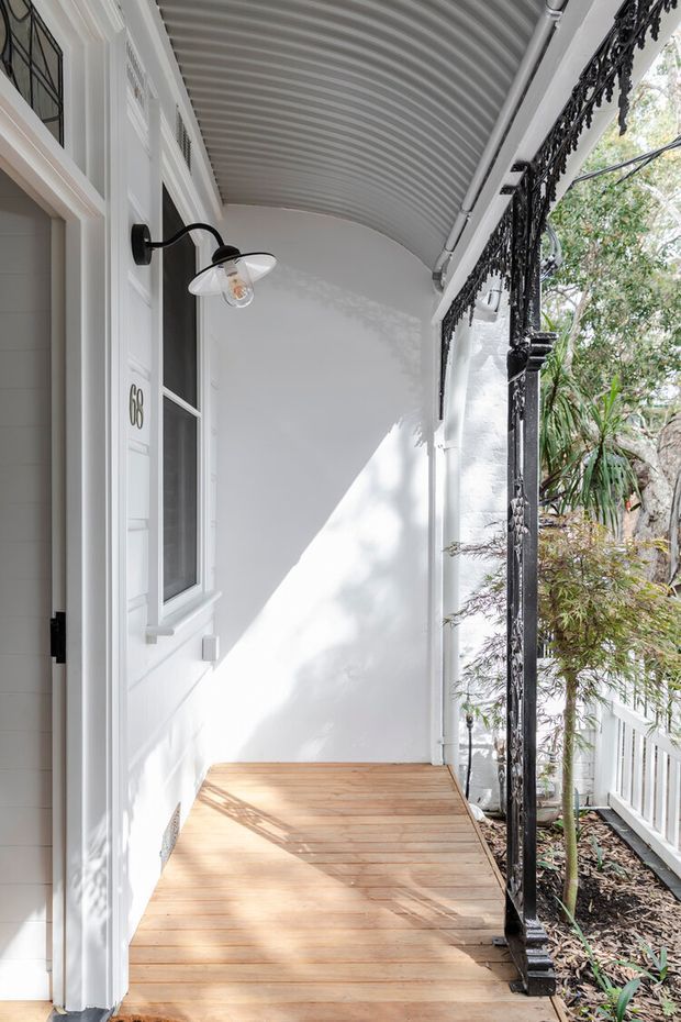 A narrow covered front verandah or porch with white painted walls, a timber entry door and window, warm timber decking, a curved ribbed ceiling, and ornate black wrought-iron posts and trim. A wall-mounted exterior lantern, house number 68, white railing, and planted garden are visible. The bright scene has a refined heritage character.