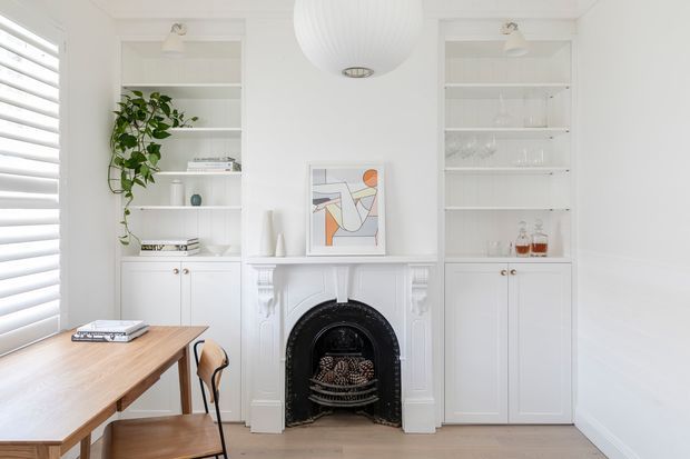 Bright, refined dining or living space with a white-painted traditional fireplace centered between symmetrical built-in display cabinetry. The room features open shelving, white lower cupboards, a timber dining table with simple chairs, a large white pendant light, plantation shutters, and pale timber flooring. An abstract artwork and restrained styling add a contemporary touch to the classic architectural detailing.