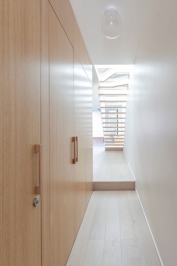 Bright minimalist interior hallway with full-height light timber built-in cabinetry and concealed doors along the left wall, pale timber flooring, white walls and ceiling, and a clear glass pendant light. The corridor leads to a short step and an open-riser timber staircase with a skylit or clerestory-lit void beyond. The space is clean, linear and naturally illuminated.