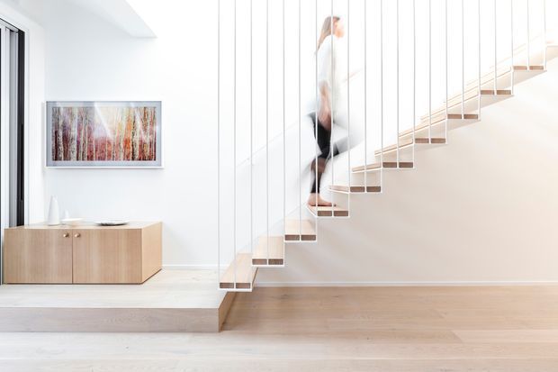 Bright minimalist interior hallway and stairway with a floating timber staircase suspended by slender vertical metal rods and a glass or open balustrade. Pale timber flooring, white walls, a raised platform, and streamlined light-wood built-in cabinetry create a clean contemporary setting. A framed artwork and a partially visible person appear in the background.