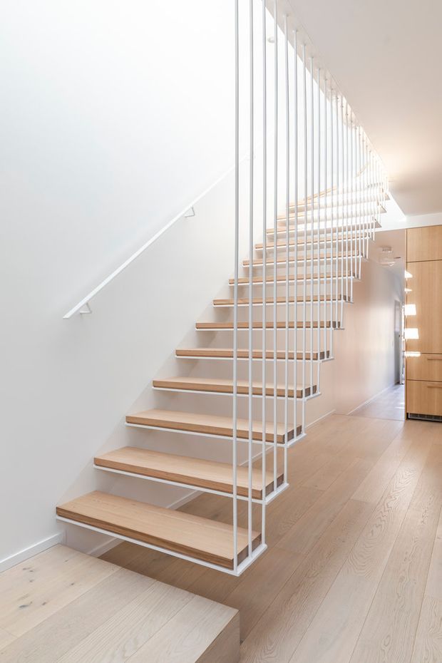 Bright contemporary interior focused on an open-riser staircase with pale timber treads, white stringers, and a distinctive white vertical-rod balustrade extending up to the upper level. Pale timber flooring continues through the hallway, with white walls and minimalist light-wood built-in cabinetry visible at right.
