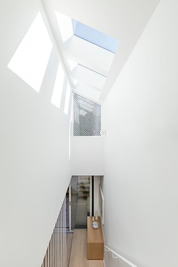 A bright, minimalist interior view looking down a narrow multi-level stairway or landing. White walls and ceiling planes surround a central void with strong angular daylight, skylight openings, and a patterned privacy screen. A light timber floor, slim white stair balustrade, and simple timber console with a round decorative object are visible below. The composition emphasises vertical space, geometric shadows, and abundant natural light.