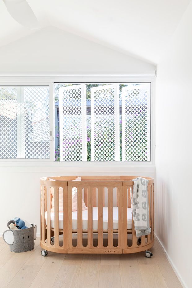 Bright, compact nursery with a rounded natural timber cot on casters positioned beneath a large white-framed window. White diamond-pattern privacy screens are visible outside the glazing, while a soft toy basket and draped baby blanket add modest furnishings. The room has white walls, a sloping ceiling, and pale timber flooring, creating an airy and clean appearance.