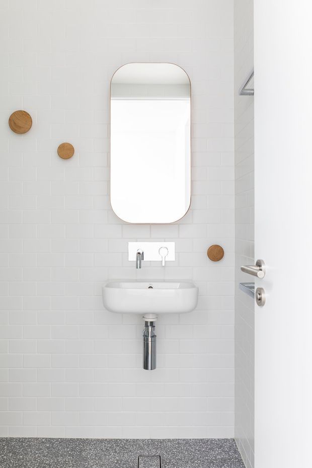 Compact minimalist bathroom with white subway-tile walls, a wall-mounted white basin, chrome exposed plumbing and tapware, and a rounded-rectangle mirror. Small circular timber wall accents add warmth, while the terrazzo-style grey floor provides contrast. A partially open white door and shower detail are visible at right.