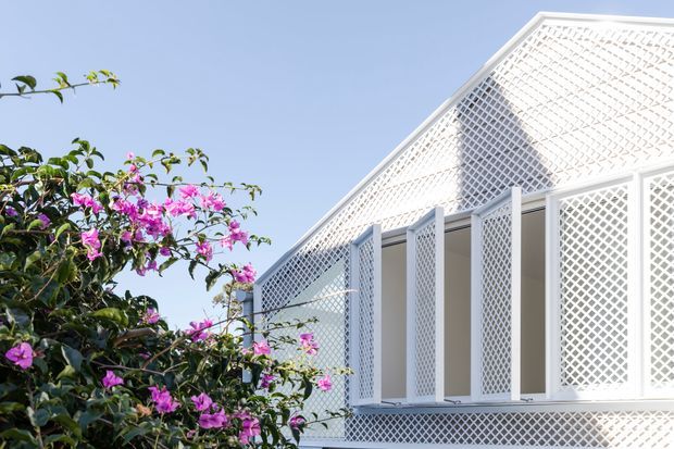 Close-up exterior view of a bright white building facade with a steep gabled roof and extensive diamond-pattern lattice screening over the upper gable, windows, and lower wall. Pink flowering vegetation partially frames the left side against a clear blue sky.
