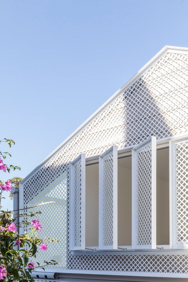 Close-up exterior view of a bright white gabled building facade with extensive diamond lattice screens, projecting vertical supports, and an open verandah or balcony. Pink flowering vegetation appears at the left edge against a clear blue sky. The image emphasizes the decorative geometric screening and light coastal character of the facade.