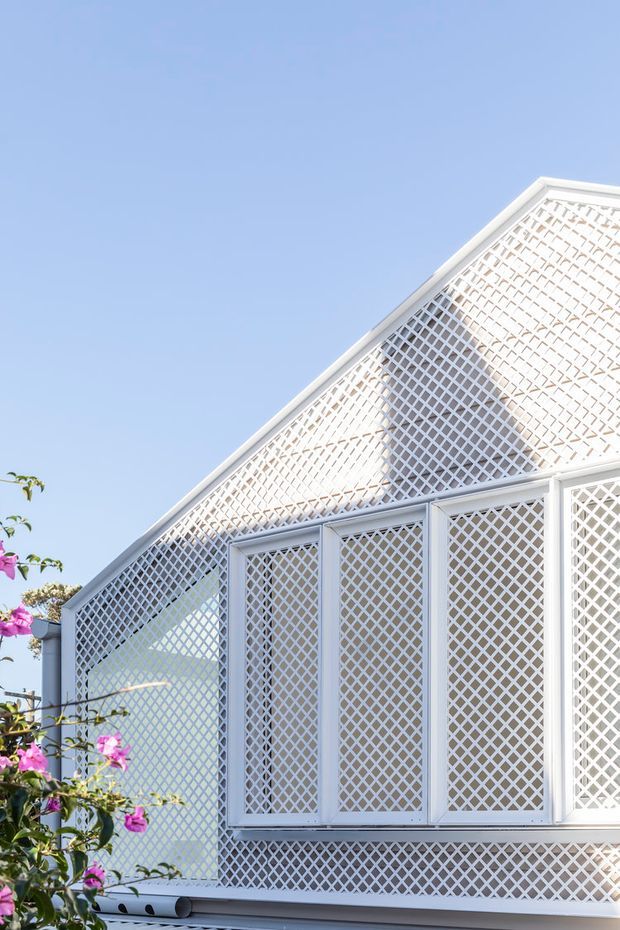 Exterior close-up of a white gabled building facade with extensive diamond-pattern lattice screens covering the gable and windows. The bright, lightweight facade is photographed against a clear blue sky, with flowering bougainvillea at the edge of the frame.