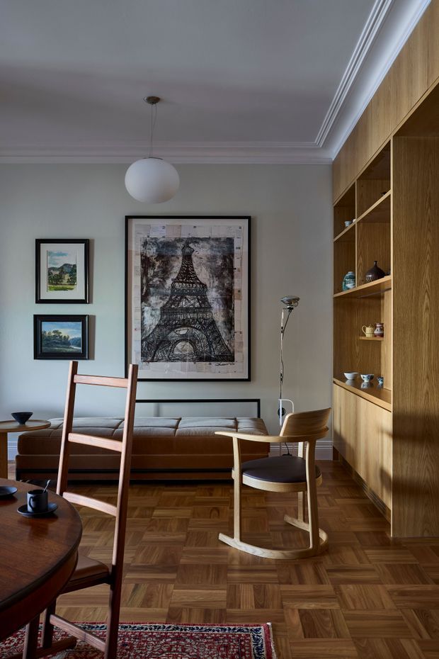 A refined mid-century-inspired living and dining area with a warm timber parquet floor, built-in timber shelving, and a brown leather sofa. The room has pale grey walls with decorative ceiling moulding, a white globe pendant, framed artwork, a sculptural timber rocking chair, and a partially visible round dining table with leather seating. The composition is warm, curated, and richly layered with timber and vintage furnishings.