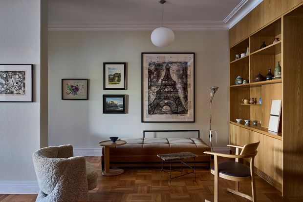A refined living room with a warm mid-century-inspired interior. The room features a brown leather sofa, textured boucle armchair, timber parquet flooring, pale grey walls with detailed cornices, framed artwork, a spherical pendant light, a slim floor lamp, and extensive built-in timber shelving and cabinetry along the right wall. A timber-framed chair and glass-topped coffee table complete the arrangement.