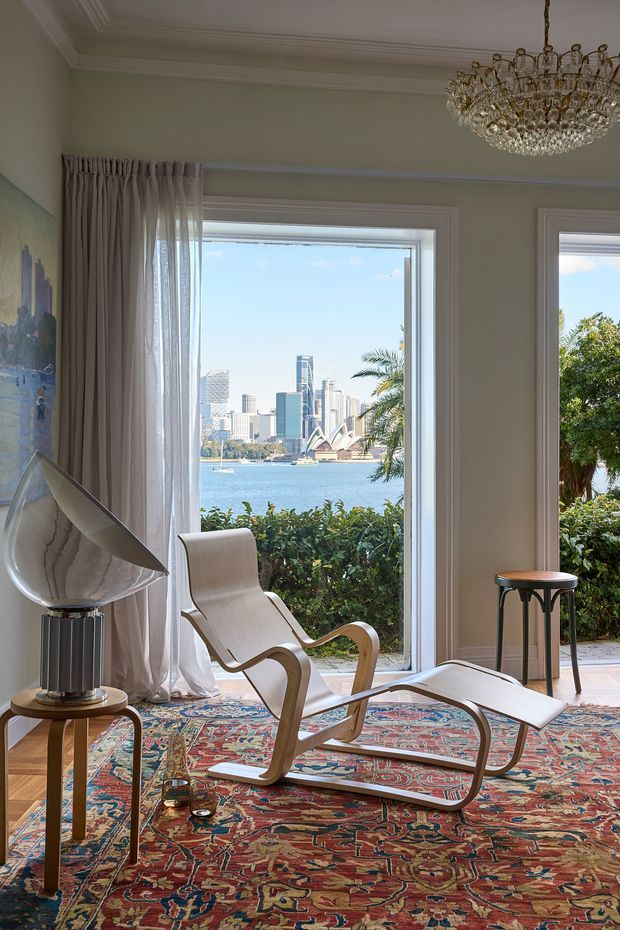 A refined sitting room with a dramatic harbour and city view through tall windows and glazed doors. The room features pale green-grey walls, ornate white ceiling cornices, sheer full-length curtains, a sculptural mid-century-style lounge chair and ottoman, a marble-and-metal table lamp, small side tables, and a richly patterned Persian-style rug. Sydney Opera House and the surrounding city skyline are visible beyond the water.