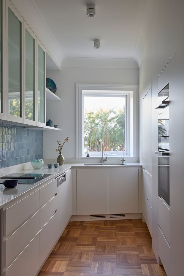 A compact, bright galley kitchen with white built-in cabinetry, full-height appliance cabinetry, pale stone benchtops, a blue-grey glazed tile splashback, and a timber parquet floor. A large window above the sink brings in daylight and views of palms, while open shelving and glass-fronted upper cabinets provide display storage. The room has a clean, refined contemporary feel with subtle traditional detailing around the window and ceiling.