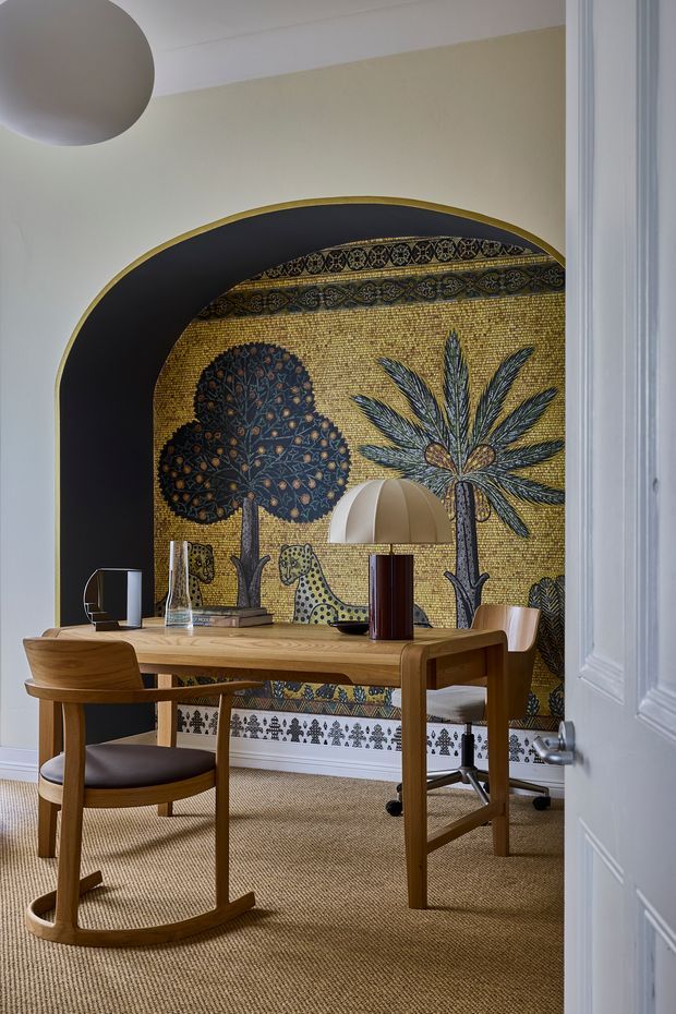 A refined dining or workspace nook viewed through a partially open white paneled door. A warm timber table is surrounded by wooden chairs, including a rocking chair, with a desk chair at the far side. The room features a dramatic arched black alcove with a gold-edged border and a large decorative mosaic mural depicting stylised trees, foliage and an animal. A dome-shaped table lamp, glassware and small objects sit on the table. The space has pale warm walls, a woven neutral carpet, and a strong decorative, jewel-toned focal wall.