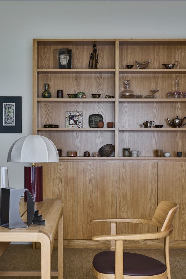 A refined interior vignette featuring a full-height built-in timber display and storage wall with open shelves filled with ceramics and decorative objects above closed lower cabinetry. In front is a light timber desk or console with a sculptural white dome lamp, burgundy base, and dark monitor, alongside a light timber armchair with a dark upholstered seat. The room has a muted grey wall, warm wood tones, and a textured neutral carpet, creating a curated mid-century-inspired study or living space.