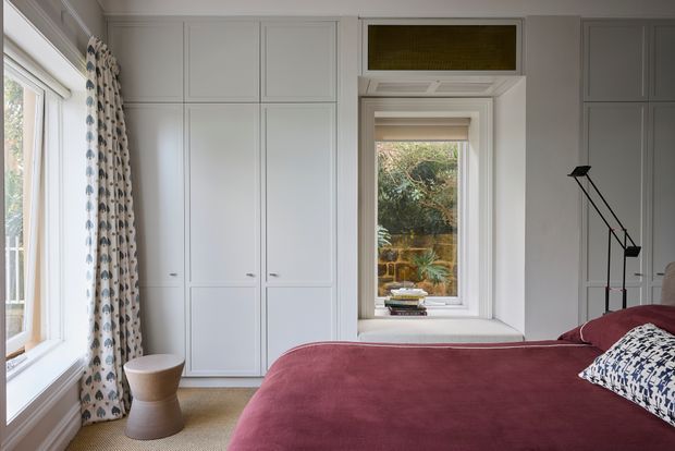 A bright, refined bedroom with white built-in wardrobes, a deep burgundy bedspread, patterned curtains and cushions, and large windows overlooking greenery. The room features classic panelled cabinetry and trim, a compact sculptural side table, and a slim black bedside lamp.